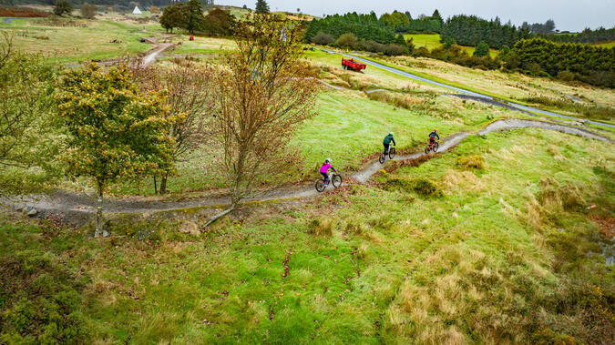 Flera organisationer inom projektet arbetar med att tillgängliggöra stigcykling för barn och unga. Foto: Beyond Horizons Flygbild över landskap med gräsängar och träddungar. Två personer syns, som cyklar mountainbike på en smal stig.